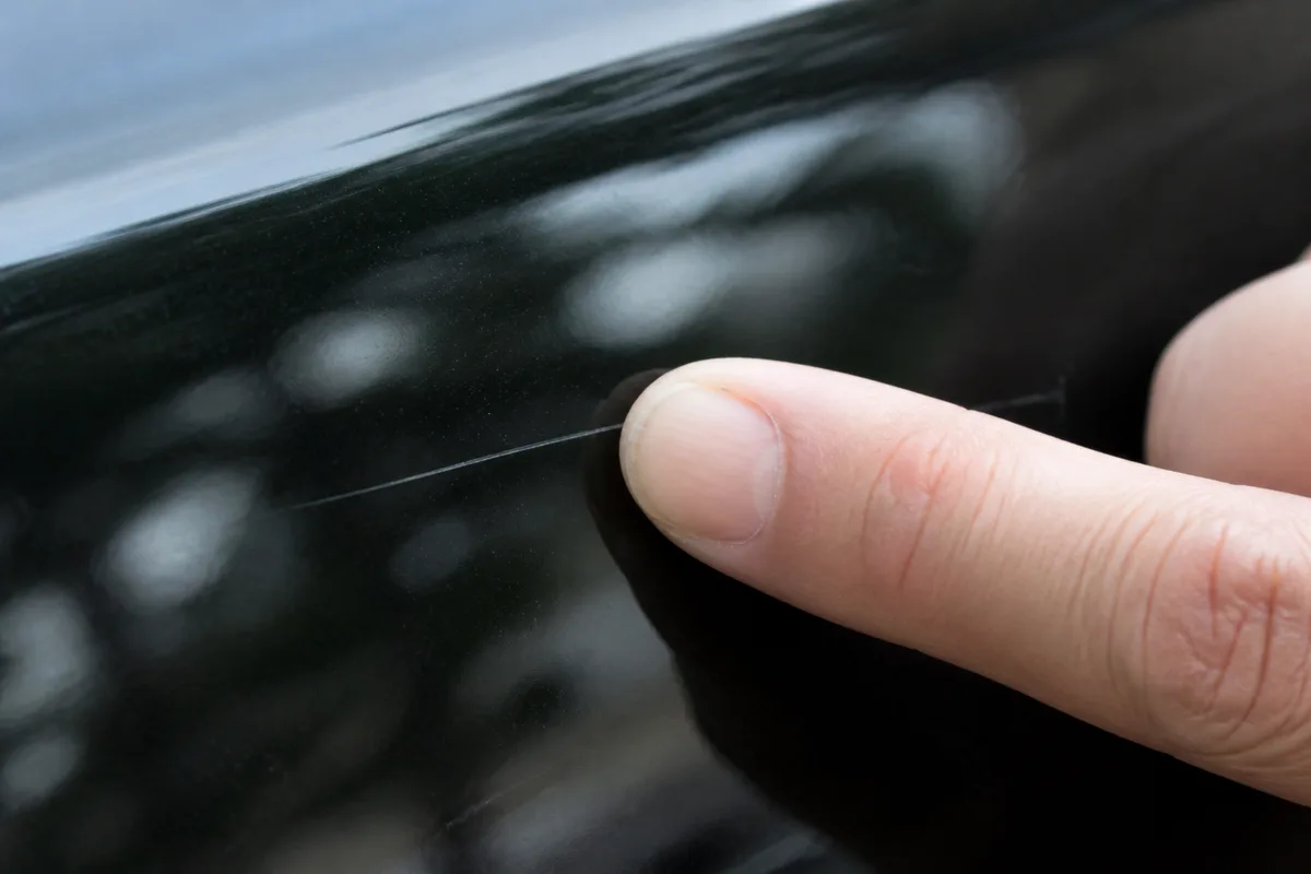 Main passant un doigt sur une rayure claire visible sur une carrosserie sombre